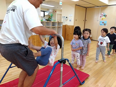 暑くて外に行けない日も室内で様々な運動遊びをしている３歳児さん。自分で足かけ回りが出来るように先生に補助してもらいながら挑戦しています！